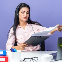 young-beautiful-woman-casual-clothes-wearing-headset-with-microphone-holding-clipboard-with-blank-pages-looking-confident-sitting-table-with-laptop-blue-wall-working-office_141793-127876